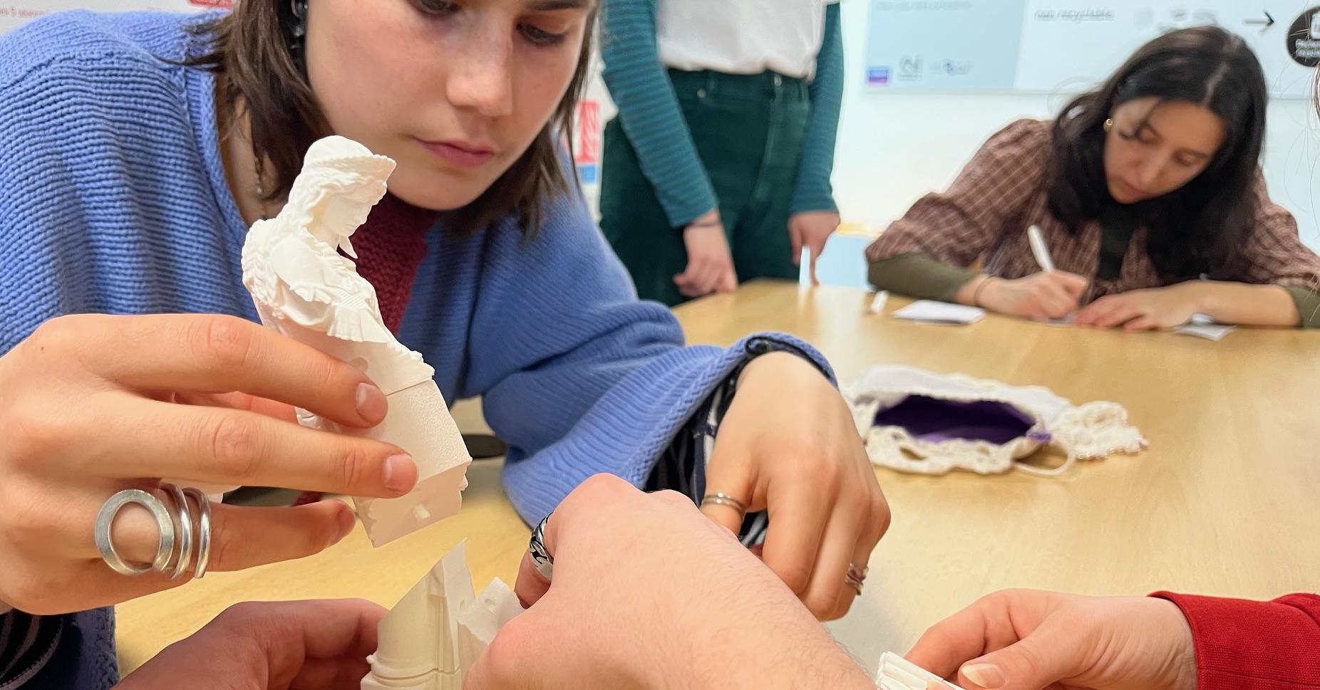 Photo de trois personnes en train d'assembler différents éléments en plastique blanc, imprimés en 3D, afin de constituer une statuette de déesse grecque d'une vingtaine de centimètres de haut.