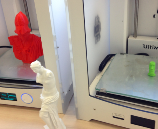 Photo des deux imprimantes 3D de l'Edulab. Dans celle à gauche de l'image, une petite statuette rouge représentant le buste d'un soldat vient d'être imprimée. Dans celle de droite, c’est la tête verte d’un bonhomme qui a été imprimée en 3D. Devant les imprimantes est disposée une reproduction d’une dizaine de centimètres de haut d’une statue grecque, représentant une femme.