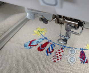Photo en gros plan de l'aiguille d'une brodeuse numérique en train de broder sur un tissu beige, des tongs en rouge, blanc et bleu, suspendues à un fil à linge, tenu par des petits soleils.