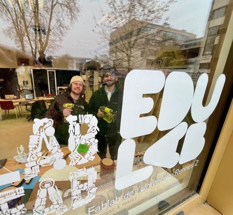 Photo d'Axel Ropert et Thomas Mouchoux tenant chacun une perceuse, derrière la vitre située à l'entrée de l'Edulab