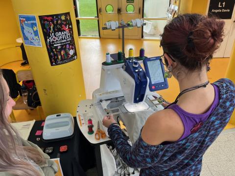 Photo de deux étudiantes en train de réaliser une broderie numérique. La machine ressemble à une machine à coudre utilisable debout, avec un écran numérique et jusqu’à quatre bobines de fils de couleurs différentes.