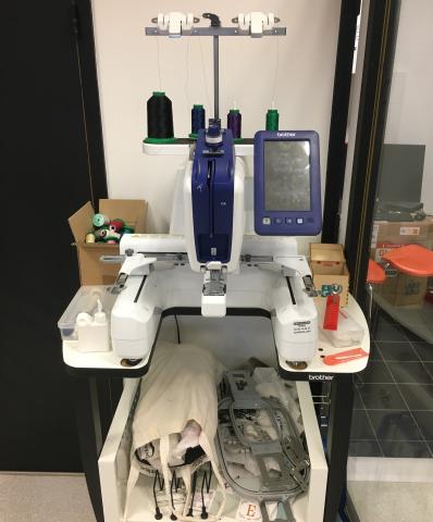 Photo de la brodeuse numérique : elle ressemble à une machine à coudre utilisable debout, avec un écran numérique et jusqu’à quatre bobines de fils de couleurs différentes.