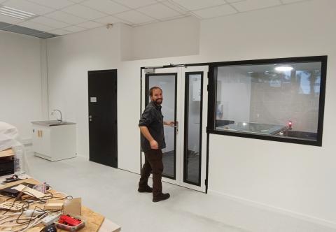Photo du fabmanager devant le nouveau sas insonorisé : une double porte vitrée et une fenêtre intérieure ont été ajoutées. La découpeuse laser a été déplacée de l’entrée de l’Edulab dans ce nouvel espace clos. La porte des toilettes a été déplacée pour s'ouvrir sur l'espace principal. L'ancienne kitchenette a été replacée dans le coin gauche de l'espace, à côté des sanitaires.
