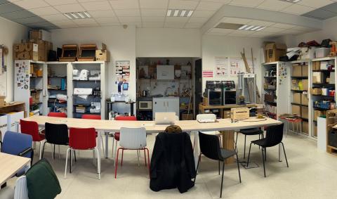 Photo de l'Edulab avant les travaux, prise depuis l'entrée : une grande table et des chaises sont disposées en travers de la pièce. Au fond de l'espace principal, une kitchenette dans une alcôve. A gauche de celle-ci, la porte des toilettes et à droite, celle du local à bois. De part et d’autres de la pièce, des armoires remplies de matériel.