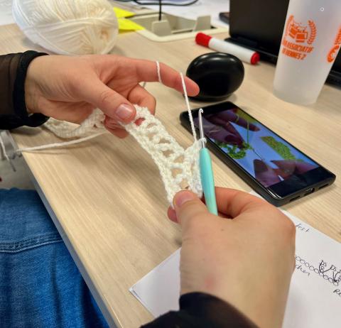 Photo des mains d'une personne en train de réaliser quelque chose au crochet, avec de la laine blanche. La personne s'aide d'une vidéo qu'elle regarde sur son téléphone.