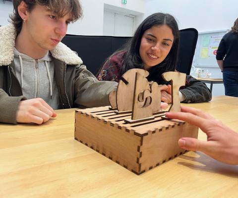 Photo d'un jeune homme et d'une jeune femme assis autour d'une table. Ils manipulent un coffret en bois sur lequel sont posées des planches de contre-plaqué, découpées en formes énigmatiques.