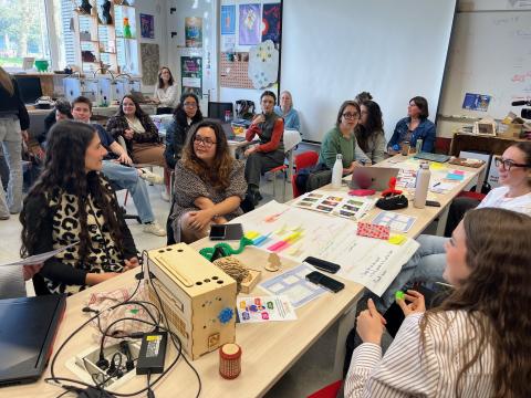 Photo représentant une vingtaine de personnes (étudiantes, étudiants, enseignantes et ingénieures pédagogiques de la DAP) réunies dans l'Edulab. Elles sont pour la plupart assises autour de tables, sur lesquelles sont disposés des documents et des exemples d'objets réalisés à l'Edulab. En arrière-plan, un tableau blanc, un écran blanc de vidéo-projection et des affiches accrochées au mur.