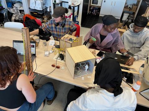 Photo représentant cinq étudiantes et étudiants de 3e année de licence, assis autour d'une table sur laquelle sont disposés les projets artistiques de fin d'année qu'elles et ils sont en train de concevoir. En arrière-plan, des armoires remplies de matériel, des brodeuses numériques utilisées à l'Edulab.