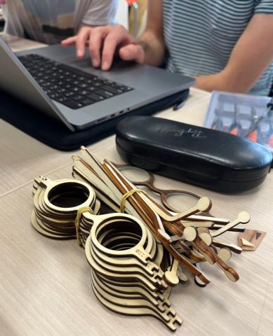 Photo d’une dizaine de montures de lunettes en contreplaqué, de tailles différentes, empilées sur une table. En arrière-plan, un boitier à lunettes noir, ainsi qu’un ordinateur sur lequel deux personnes sont en train de travailler.
