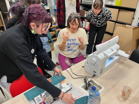 Photo de cinq personnes réunies autour d'une table sur laquelle est posée une brodeuse numérique. En arrière-plan, des armoires remplies de cartons et de matériel utilisé à l'Edulab.