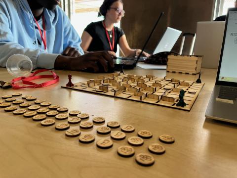 Photo d'une table sur laquelle est posé un labyrinthe et des pions en contreplaqué, réalisés grâce à la découpeuse laser de l'Edulab. En arrière-plan, un étudiant et une étudiante en train de travailler sur ordinateurs.