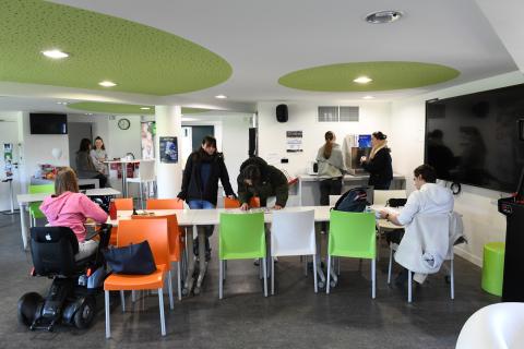 Photo de la salle de convivialité du Ty-Maz'. Au premier plan, 3 tables et des chaises blanches, orange et vertes. Une étudiante en fauteuil est en train de travailler sur son ordinateur, deux autres debout lisent un document, un 4e est en train de lire un document.  Un très grand écran vidéo est fixé au mur de droite. A l’arrière-plan, deux étudiantes discutent devant un distributeur d’eau et un micro-ondes. Tout au fond, deux autres étudiantes discutent autour d’une table « mange-debout ».