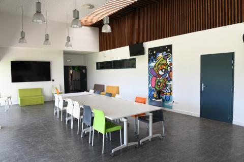 Photo de la salle de pédagogique et culturelle du Ty-Maz'. Au premier plan, une grande table autour de laquelle sous disposées douze chaises blanches, grises, orange et verte. A l’arrière-plan, trois fauteuils vert, jaune et gris, ainsi qu’un très grand écran vidéo noir fixé au mur.