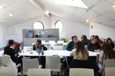 Photo de la salle d'étude du Ty-Maz'. L’espace est lumineux grâce à deux fenêtres hublots et des velux au plafond. Au premier plan, des tables et chaises blanches autour desquelles une dizaine d’étudiantes et étudiants sont en train de travailler. A l’arrière-plan, un meuble gris où sont disposés un distributeur à eau et deux micro-ondes.