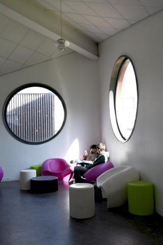 Espace libre du Ty-Maz' où deux étudiants sont en train de discuter, assis dans des fauteuils arrondis et colorés. La lumière de l'espace est douce, grâce à deux fenêtres hublots, situées juste au-dessus des fauteuils.