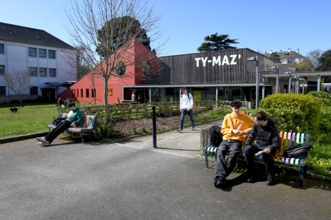 Photo du Ty-Maz' vu de l'extérieur. Au premier plan, de chaque côté d'une allée, deux bancs en métal peints aux couleurs de l'arc-en-ciel, sur lesquels sont assis des étudiants. Un 4e étudiant venant du Ty-Maz' marche dans l'allée. A l'arrière-plan, le bâtiment du Ty-Maz' dont la partie gauche est rouge, construite en triangle, avec des fenêtes hublots et la partie droite est rectangulaire, plane et recouverte d'un bardage en bois.