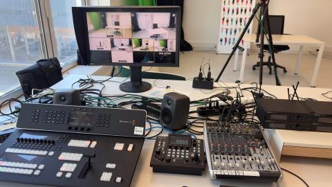 Photographie du studio audiovisuel représentant au premier plan un écran d'ordinateur, des enceintes et trois tables de mixage. En arrière-plan, une caméra sur pied.