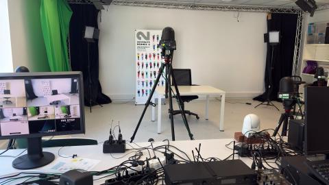 Photographie du studio audiovisuel représentant une caméra sur pied et en arrière-plan : des spots lumineux éteints, des rideaux permettant de faire un fond noir ou vert.