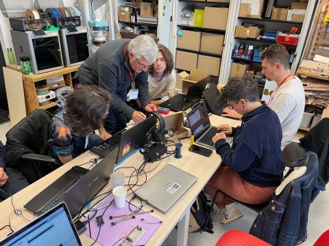 Photo prise dans l'Edulab : quatre personnes sont assises devant leurs ordinateurs ouverts autour d'une table. Un homme est debout et semble réaliser des tests électriques sur un petit appareil. A droite de l'image, des placards sont ouverts et remplis de cartons et boîtes de rangement. Au fond, deux imprimantes numériques.