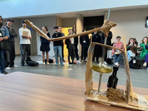 Photo prise dans le hall du Pôle numérique Rennes Villejean (PNRV), situé au bâtiment T. Au premier plan, un prototype en carton et métal d'une dynamo permettant de recharger un téléphone portable. A l'arrière, une quinzaine de personnes assistant à la présentation du projet.