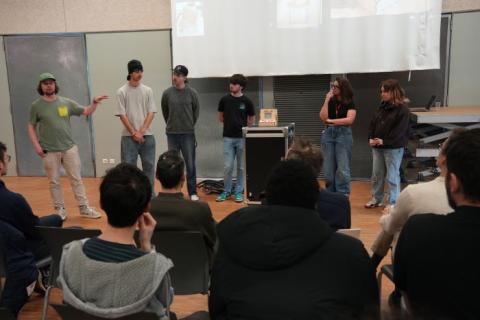 Photo de six personnes debout présentant une mini borne d'arcades en bois, devant un public assis.